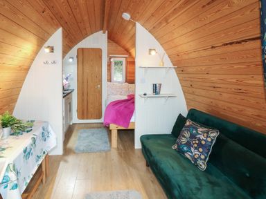 A small cabin interior with wooden walls featuring a green sofa with a patterned pillow a dining table with a floral tablecloth a bed with a pink blanket and a wooden door in the back at Chantelles in Holbeach