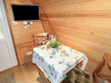 A small dining area with a table covered by a floral tablecloth two wooden chairs a bottle of wine two wine glasses a metal ice bucket a small plant a wall-mounted flat screen television and wooden panel walls at Chantelles in Holbeach