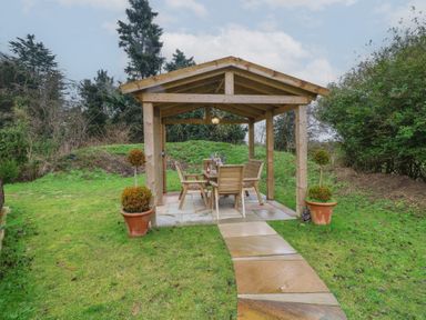 A wooden gazebo with a table and chairs inside on a stone patio in a garden with potted plants and a stone path leading to it at Chantelles in Holbeach