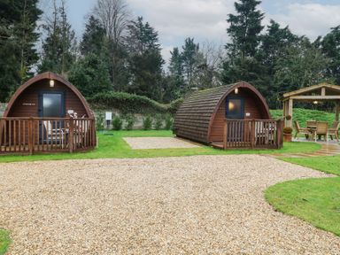 Two wooden camping pods with small decks in a grassy area near a covered outdoor seating area surrounded by trees at Chantelles in Holbeach