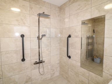 A tiled shower area with a shower head and handheld shower and two wall grab bars with a mirror reflecting a mop and bucket at Chantelles in Holbeach