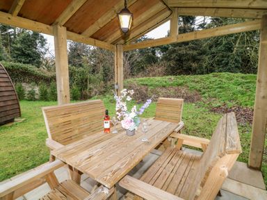 A wooden outdoor seating area with a table, benches, chairs, a bottle of wine, glasses, and a flower arrangement at Charlottes in Holbeach