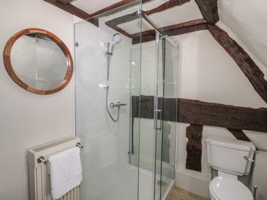 A bathroom with a glass shower enclosure a toilet a round wall mirror and wooden beams at Church View in Welford-On-Avon