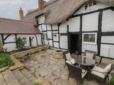 An outdoor patio with stone flooring a round table with four chairs a stone bench flowerbeds and a thatched roof cottage at Church View in Welford-On-Avon