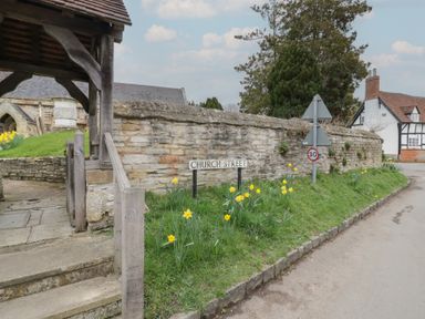 A stone wall with a church street sign and yellow flowers along the roadside near a wooden gate and a half-timbered house at Church View in Welford-On-Avon