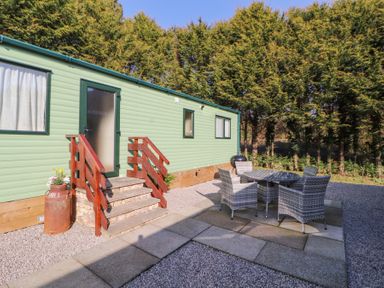 An outdoor patio area with wicker chairs and a round table next to a green mobile home with wooden steps and trees in the background at Lodge 1 in Silverdale