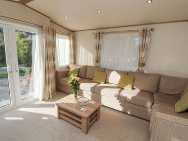 A sitting room with a beige corner sofa green cushions wooden coffee table with flowers and large windows with curtains at Lodge 1 in Silverdale