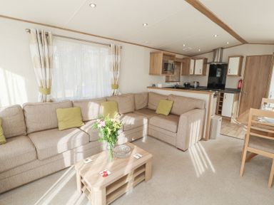 A living room with a beige sectional sofa and green cushions a wooden coffee table with flowers and coasters a kitchen area with cabinets and appliances in the background at Lodge 1 in Silverdale