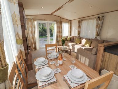 A dining table set with plates bowls glasses and a bottle next to a sofa and windows with curtains at Lodge 1 in Silverdale