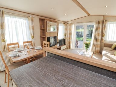 A living room with a dining table set for four a sofa a coffee table with flowers a TV and large windows with curtains at Lodge 1 in Silverdale