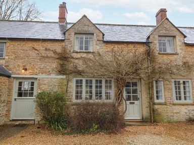 A stone cottage with multiple windows a vine climbing around the door and bushes in front at Little Lemhill Farm Cottage in Lechlade-On-Thames