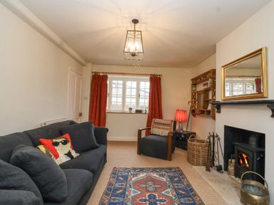 A living room with a black sofa with decorative pillows an armchair a patterned rug a fireplace with a wood-burning stove and a window with red curtains at Little Lemhill Farm Cottage in Lechlade-On-Thames
