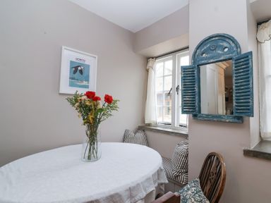A dining area with a round table covered with a white cloth and a vase of flowers a blue shutter-style mirror on the wall a window with white curtains and striped cushions on a window seat at Little Lemhill Farm Cottage Lechlade-On-Thames