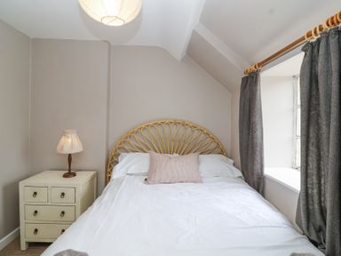 A bedroom with a bed with a rattan headboard a side table with a lamp and a window with curtains at Little Lemhill Farm Cottage in Lechlade-On-Thames