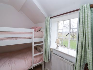 A bedroom with white bunk beds next to a window with green curtains and a small lamp on the windowsill at Little Lemhill Farm Cottage in Lechlade-On-Thames