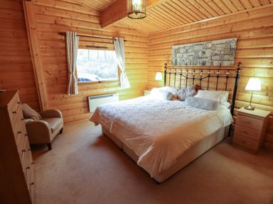 A bedroom with a double bed two bedside tables lamps a chest of drawers an armchair and a window in a wooden cabin at Forest Lodge in Kenwick Woods