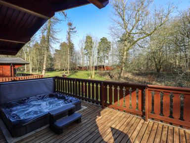 An outdoor wooden deck with a hot tub and railing overlooking trees and grass at Forest Lodge in Kenwick Woods