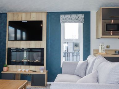 A living room with a mounted television above an electric fireplace a window with a floral roman blind and a sofa with cushions at The Maples No 48 in Hewas Water near Sticker