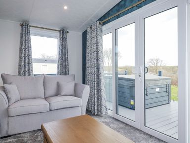 A living room with a gray sofa a wooden table and large sliding glass doors leading to a deck with a hot tub at The Maples No 48 in Hewas Water near Sticker