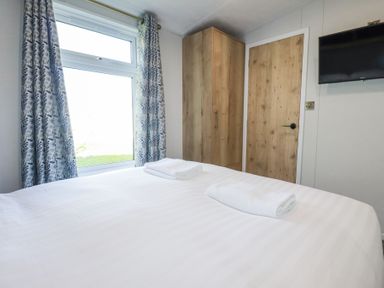 A bedroom with a bed covered in white linens two folded towels on the bed a window with patterned curtains a wooden wardrobe a wooden door and a wall-mounted television at The Maples No 48 in Hewas Water near Sticker