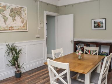 A dining room with a wooden table and four white chairs a framed world map and a photo of a cow on the wall a tall plant in a pot and a bookshelf with books at Heatherbell House in Ballyhoura near Buttevant