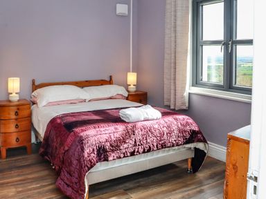 A bedroom with a bed covered with a purple blanket two bedside tables with lamps and a window showing a view at Heatherbell House Ballyhoura near Buttevant