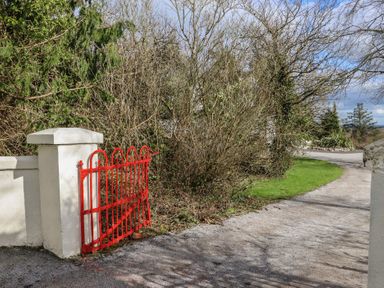 A red metal gate attached to a white wall with a gravel driveway and trees and bushes at Heatherbell House in Ballyhoura near Buttevant