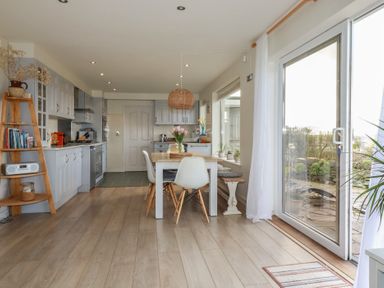A kitchen and dining area with a wooden table white chairs sliding glass door and wooden shelves at Coastmans Nest in Mawgan Porth