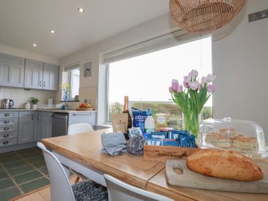 A kitchen with grey cabinets a wooden dining table with flowers bread and snacks at Coastmans Nest in Mawgan Porth