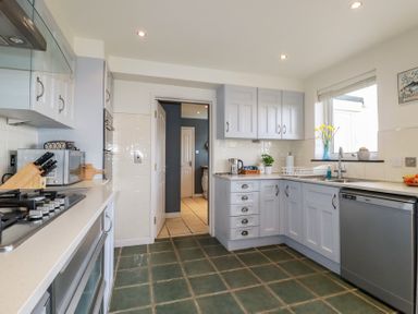 A kitchen with white cabinets a gas stove a dishwasher and a window with flowers at Coastman's Nest in Mawgan Porth