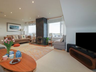 A living room with a brown sofa, a gray armchair, a wooden coffee table with a tea set and flowers, a fireplace, and a television at Coastman's Nest in Mawgan Porth