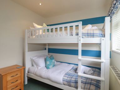 A bedroom with white bunk beds blue and white bedding a wooden bedside table and a window with blinds at Coastman's Nest in Mawgan Porth