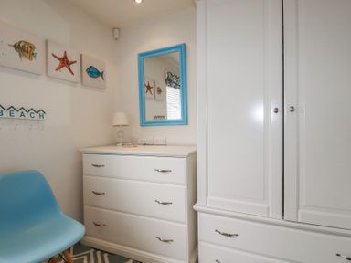 A room with a white chest of drawers a white wardrobe a blue framed mirror a blue chair and coastal themed wall art at Coastman's Nest in Mawgan Porth