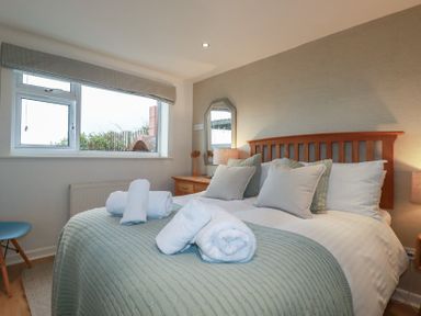 A bedroom with a wooden bed frame four pillows rolled towels on a light green blanket bedside tables with lamps and a window at Coastman's Nest in Mawgan Porth