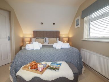 A bedroom with a bed, bedside tables with lamps, a window with blinds, towels on the bed, and breakfast tray with croissants and coffee at Coastman's Nest in Mawgan Porth