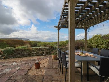 An outdoor patio dining area with table and chairs under a wooden pergola with string lights at Coastmans Nest in Mawgan Porth
