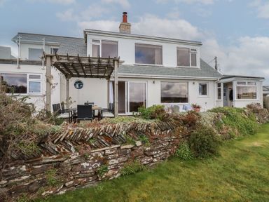 The back of a house with a wooden pergola over an outdoor dining area and a stone wall in a garden at Coastman's Nest in Mawgan Porth