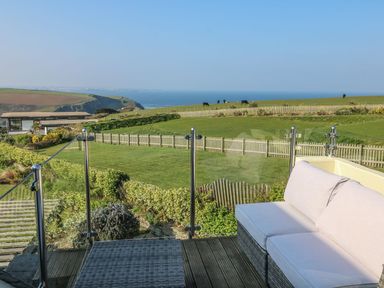 A balcony with wicker sofa and low table overlooking green fields and a sea view at Coastmans Nest in Mawgan Porth
