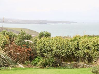 A garden with shrubs and grass overlooking a body of water and distant hills at Coastman's Nest in Mawgan Porth
