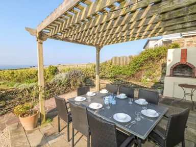 An outdoor dining area with a table and chairs under a wooden pergola with a built in brick barbecue and garden in the background at Coastmans Nest in Mawgan Porth