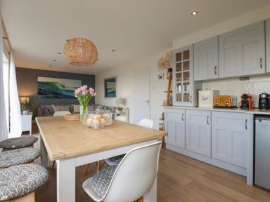 A kitchen and dining area with a wooden table and chairs a living room with a sofa and wall art at Coastman's Nest in Mawgan Porth