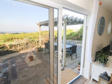 A patio area with a wooden pergola and outdoor dining table and chairs viewed through glass sliding doors at Coastmans Nest in Mawgan Porth