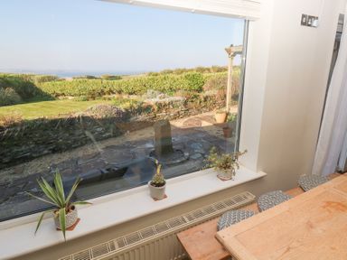 A dining area with a wooden table and three stools next to a large window overlooking a garden and fields at Coastmans Nest in Mawgan Porth