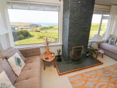 A living room with two sofas a stone fireplace a small wooden table and large windows showing a green garden and coastline at Coastman's Nest in Mawgan Porth