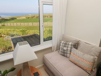 A corner with a cushioned bench seat, two patterned pillows, a small round table with a lamp, and a window showing green fields and the sea at Coastman's Nest in Mawgan Porth