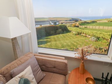 A corner of a living room with a brown sofa cushions beside a wooden table and lamp with a window showing a garden and fields at Coastman's Nest in Mawgan Porth