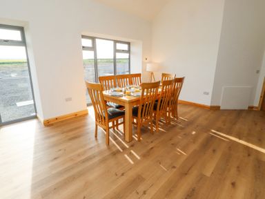 A dining area with a wooden table and eight chairs next to large windows at West Burton Hemmel in Bamburgh