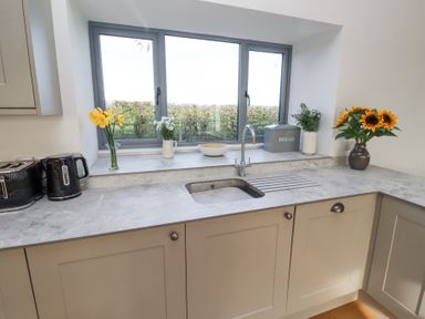 A kitchen sink under a window with flowers bread bin and black kettle on countertop at West Burton Hemmel in Bamburgh