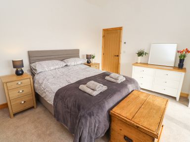 A bedroom with a bed with gray bedding and towels on top two wooden nightstands with lamps a wooden door a white dresser with a mirror and flowers and a wooden chest at the foot of the bed at West Burton Hemmel in Bamburgh