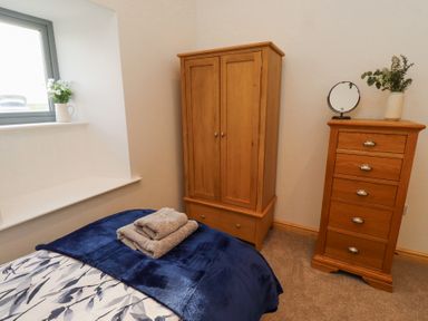 A bedroom with a wooden wardrobe a wooden chest of drawers a bed with blue and white bedding and folded towels on top and a window with a vase of flowers at West Burton Hemmel in Bamburgh